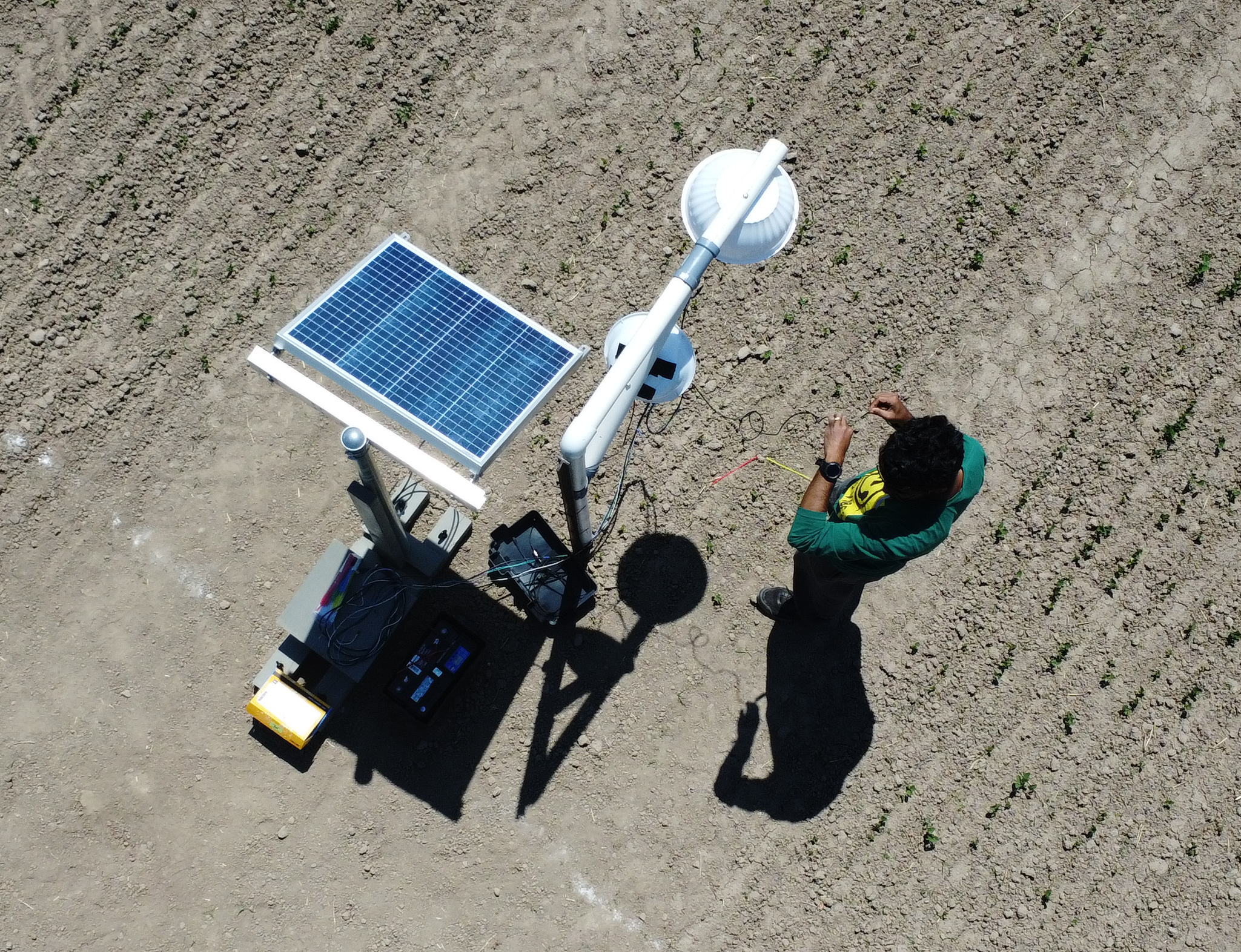 Drone image looking down at person attaching wires to a stationary camera setup including a battery, solar panel and white PVC rig with two cameras
