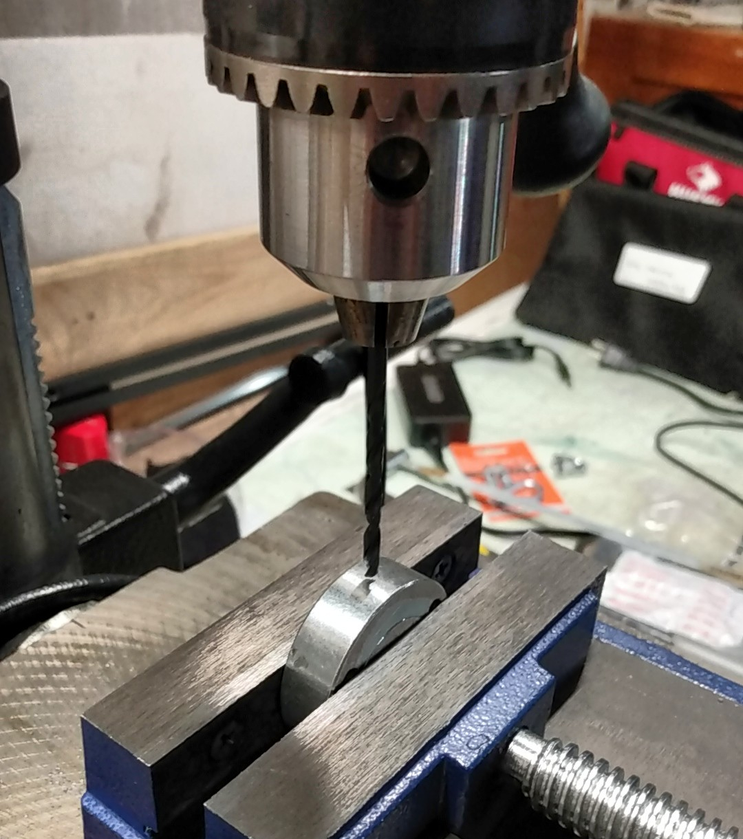 Drill press showing a drill bit just above an aluminum disk held in a vise. Out of focus junk in the background.