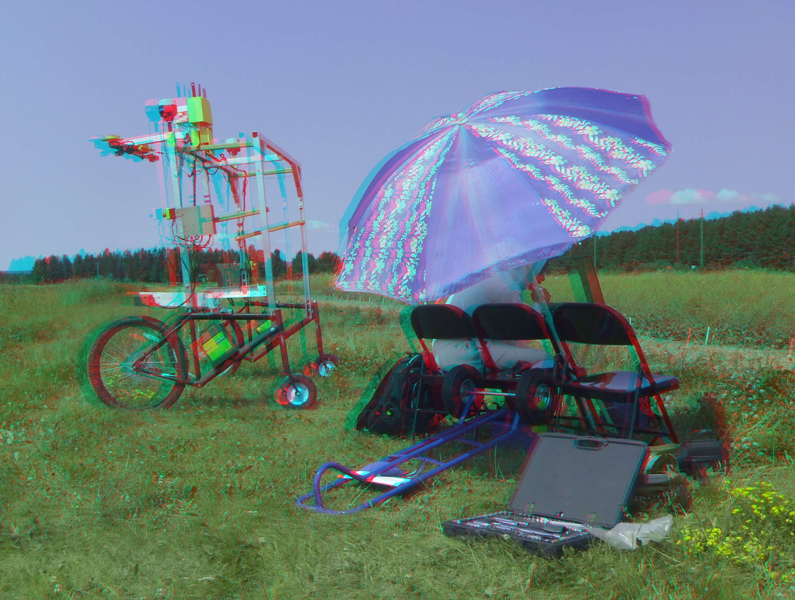 Anaglyph stereo image (red-cyan glasses) of a field work station with 3 chairs, a large umbrella, some assorted tools scattered about with a robot constructed out of used bicycle parts and aluminum uni-strut carrying multiple cameras, and fitted with 2 GPS units, a wifi router and many, many cables. This is against a background of canola, with wheat in the distance.