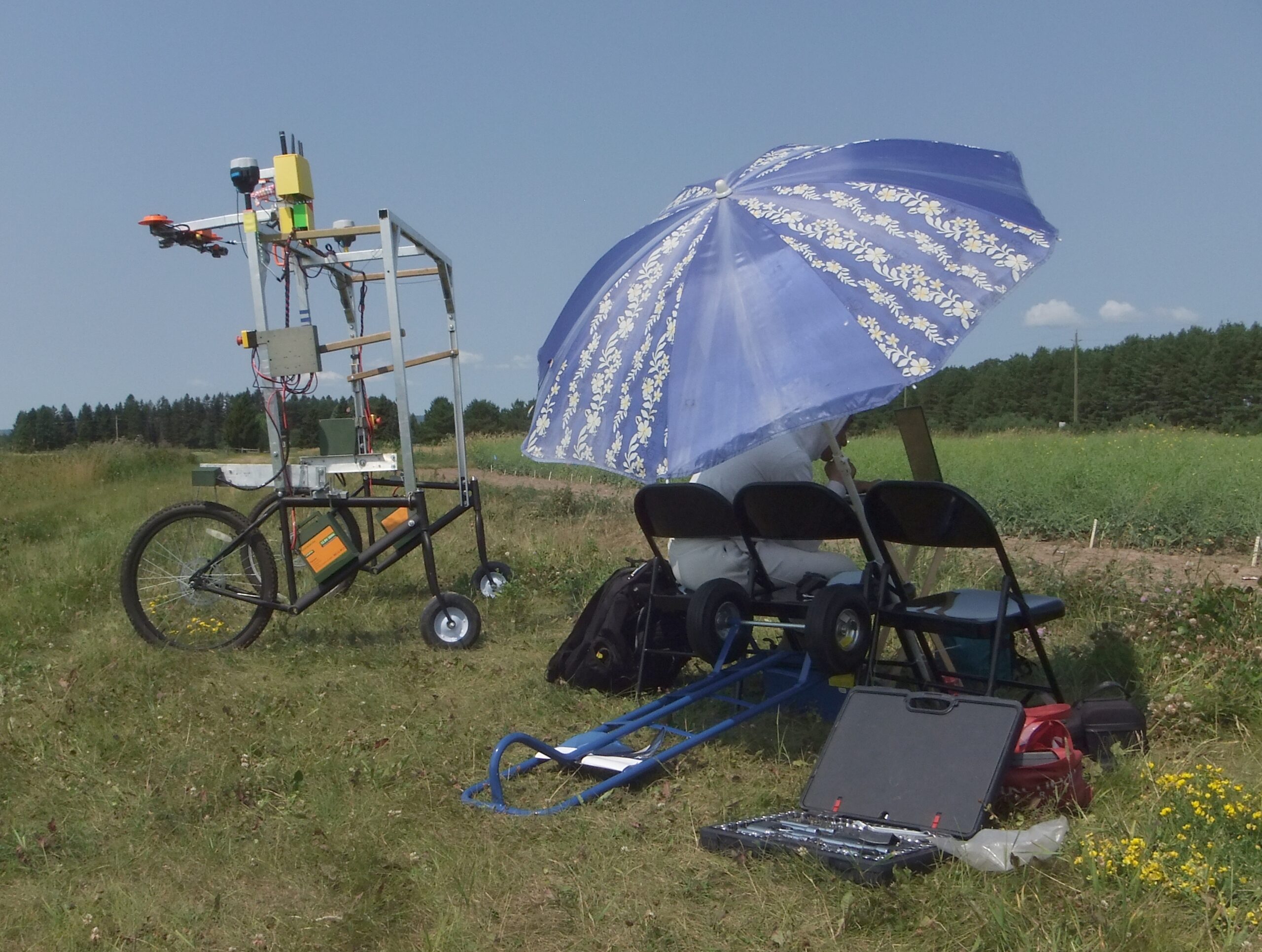 A field work station with 3 chairs, a large umbrella, some assorted tools scattered about, with a robot constructed out of used bicycle parts and aluminum uni-strut carrying multiple cameras, and fitted with 2 GPS units, a wifi router and many, many cables. This is against a background of canola, with wheat in the distance.